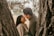 A couple is sharing a joyful and intimate moment between two tree trunks. The woman is smiling brightly as she looks at her partner. Sunlight filters through the leaves of the tree, creating a warm and tranquil atmosphere.