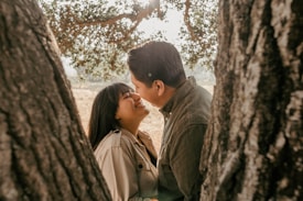 A couple is sharing a joyful and intimate moment between two tree trunks. The woman is smiling brightly as she looks at her partner. Sunlight filters through the leaves of the tree, creating a warm and tranquil atmosphere.