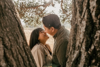 A couple is sharing a joyful and intimate moment between two tree trunks. The woman is smiling brightly as she looks at her partner. Sunlight filters through the leaves of the tree, creating a warm and tranquil atmosphere.