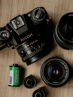 A vintage Ricoh film camera is placed on a wooden surface alongside camera lenses and a roll of Fujicolor Superia 200 film. The lenses are seen at different angles, showcasing the metallic shine, and the film has a distinct green and black casing.