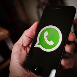 A close-up image of a hand holding a smartphone with the WhatsApp logo displayed prominently on the screen. The logo is green and white, centered against a black background.