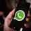 A close-up image of a hand holding a smartphone with the WhatsApp logo displayed prominently on the screen. The logo is green and white, centered against a black background.