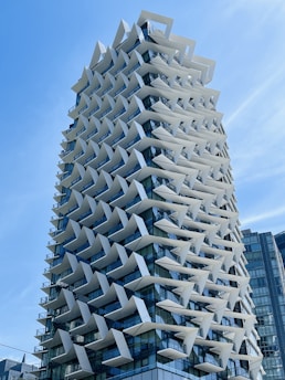 A modern high-rise building with a unique architectural design featuring numerous protruding white geometric elements. The facade consists of large glass windows lined with angled panels, creating an intricate pattern and reflecting the sky.