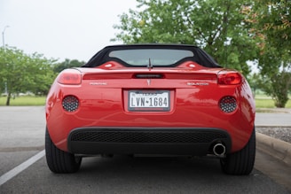 A red convertible car is parked in a lot, viewed from the rear. The vehicle is a Pontiac Solstice with a Texas license plate. Green trees are visible in the background, suggesting a natural setting.