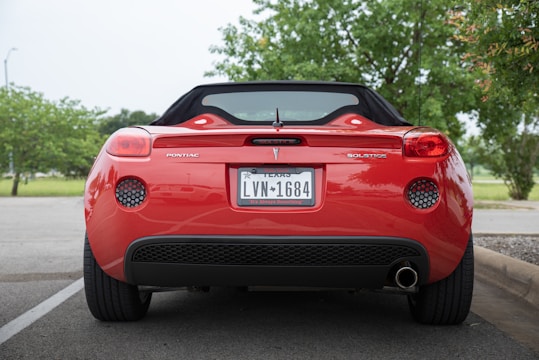 A red convertible car is parked in a lot, viewed from the rear. The vehicle is a Pontiac Solstice with a Texas license plate. Green trees are visible in the background, suggesting a natural setting.