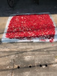 A large number of red chili peppers are spread out evenly across a rectangular cloth or mat, drying in the sun on a concrete surface. The sun casts shadows, adding contrast to the scene.
