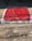 A large number of red chili peppers are spread out evenly across a rectangular cloth or mat, drying in the sun on a concrete surface. The sun casts shadows, adding contrast to the scene.