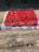 A large number of red chili peppers are spread out evenly across a rectangular cloth or mat, drying in the sun on a concrete surface. The sun casts shadows, adding contrast to the scene.