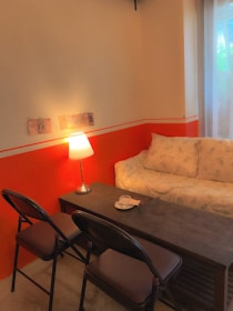 A cozy interior setting with an orange and cream color scheme. A cushioned sofa with a floral pattern is positioned against a wall, accompanied by a small wooden table with a lamp on it. Two folding chairs are placed on one side of the table. Soft natural light filters in through sheer curtains.