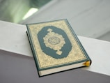 A book with an ornate gold and green cover featuring intricate patterns and Arabic script. It is placed on a light-colored surface with soft lighting in the background.