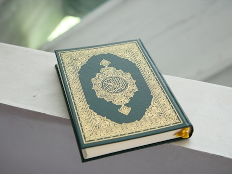 A book with an ornate gold and green cover featuring intricate patterns and Arabic script. It is placed on a light-colored surface with soft lighting in the background.