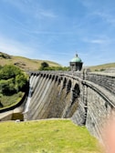 A picturesque stone dam with a series of arches holds back water, creating small waterfalls along its surface. The structure is topped with a small, ornate building that has a green, domed roof. Surrounding the dam, lush green hills and clear blue skies contribute to the serene landscape.
