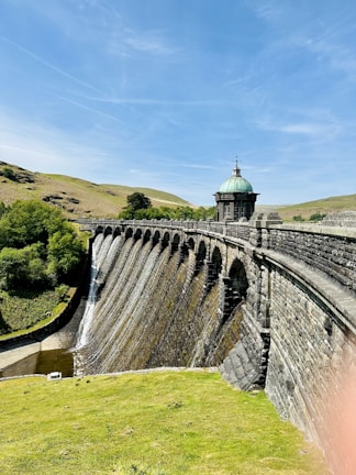A picturesque stone dam with a series of arches holds back water, creating small waterfalls along its surface. The structure is topped with a small, ornate building that has a green, domed roof. Surrounding the dam, lush green hills and clear blue skies contribute to the serene landscape.