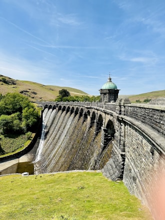 A picturesque stone dam with a series of arches holds back water, creating small waterfalls along its surface. The structure is topped with a small, ornate building that has a green, domed roof. Surrounding the dam, lush green hills and clear blue skies contribute to the serene landscape.
