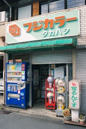 The image depicts the exterior of a small shop with a closed shutter on one side and an open entrance under a green awning. In front of the shop, there is a blue vending machine dispensing drinks and several capsule toy machines in red and gray stands. A potted plant sits near the entrance, and signage with Japanese characters is displayed prominently.