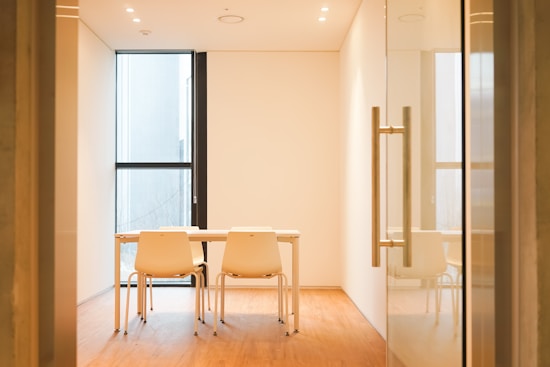 A minimalist conference room with a wooden floor, featuring a large window providing ample natural light. The room contains a light-colored table with two matching chairs set up for a meeting. Recessed ceiling lights provide additional lighting. A glass door with a metallic handle is partially visible in the foreground.