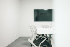 A minimalist meeting room featuring a large black screen mounted on a stark white wall. Several modern white chairs surround a clean rectangular table, with cables and electronic devices placed on the table. The room has a bright, uncluttered appearance, suggesting a professional setting.