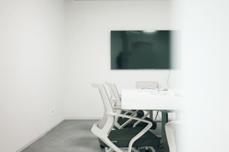 A minimalist meeting room featuring a large black screen mounted on a stark white wall. Several modern white chairs surround a clean rectangular table, with cables and electronic devices placed on the table. The room has a bright, uncluttered appearance, suggesting a professional setting.