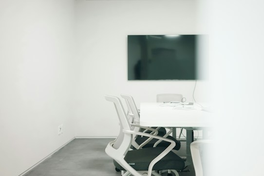 A minimalist meeting room featuring a large black screen mounted on a stark white wall. Several modern white chairs surround a clean rectangular table, with cables and electronic devices placed on the table. The room has a bright, uncluttered appearance, suggesting a professional setting.
