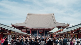 A large crowd of people gathers in front of a traditional Japanese temple. The architecture features a prominent sloping roof and intricate details. The scene is bustling with activity, with people walking around and visiting various stalls on either side of the pathway leading to the temple.