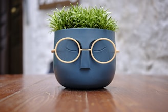 A decorative plant pot with a face design, featuring a pair of round eyeglasses and a simplistic facial expression, sits on a wooden surface. The pot holds green foliage resembling grass.