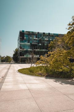 A modern building with a reflective glass facade, featuring a mix of transparent and opaque sections, is surrounded by lush green trees and landscaping. The scene is bathed in bright daylight, with a clear blue sky overhead. The foreground includes a wide, clean concrete walkway leading up to the structure.