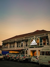 A two-story building with a traditional architectural style features a prominent adidas logo. Several cars are parked in front of the building. The sky in the background is a gradient of soft colors suggesting evening or early morning lighting.