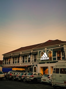 A two-story building with a traditional architectural style features a prominent adidas logo. Several cars are parked in front of the building. The sky in the background is a gradient of soft colors suggesting evening or early morning lighting.
