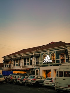 A two-story building with a traditional architectural style features a prominent adidas logo. Several cars are parked in front of the building. The sky in the background is a gradient of soft colors suggesting evening or early morning lighting.