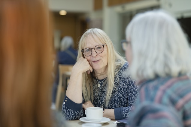 A group of older individuals sitting together at a table, engaging in conversation. One person with long hair and glasses is in focus, smiling gently while leaning on their hand. The atmosphere appears relaxed, and there are cups on the table.