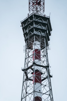 A tall metal telecommunications tower with a combination of red and white paint. The structure features multiple platforms and ladders, indicating it might be used for communication purposes. The background is a cloudy sky, suggesting an outdoor setting.