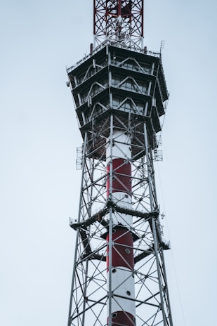 A tall metal telecommunications tower with a combination of red and white paint. The structure features multiple platforms and ladders, indicating it might be used for communication purposes. The background is a cloudy sky, suggesting an outdoor setting.