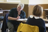 A man and woman are sitting in a classroom or meeting room. The man is smiling and holding a pen, appearing to be engaged in conversation or taking notes. The room has wooden desks and chairs with warm lighting from the windows.