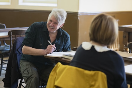 A man and woman are sitting in a classroom or meeting room. The man is smiling and holding a pen, appearing to be engaged in conversation or taking notes. The room has wooden desks and chairs with warm lighting from the windows.