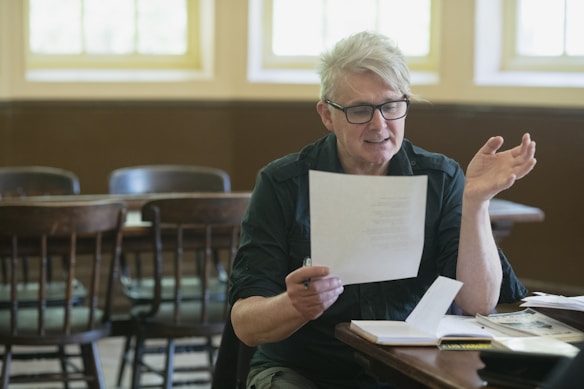 A person with glasses is sitting at a table, holding a piece of paper and gesturing with one hand. The setting appears to be a classroom or lecture room with wooden chairs and warm lighting. Various papers and books are spread on the table.