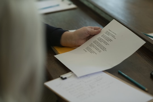 A hand holding a sheet of paper with text printed on it, possibly a poem or a piece of prose. The paper is over a notebook on a wooden surface. A pencil and another clipboard with paper are also visible.
