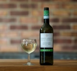 A bottle of Italian Pinot Grigio wine is positioned next to a glass filled with white wine, standing on a wooden surface. The label on the bottle is clearly visible and provides specific details about the wine. The background is a blurred brick wall, adding a rustic ambiance to the scene.