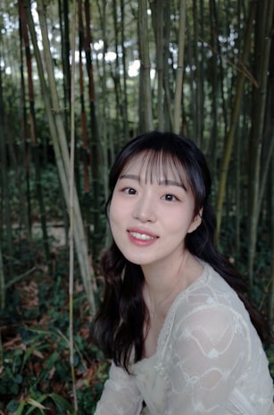 A person with long dark hair and a delicate lace top is seated amidst a dense bamboo grove. The background consists of tall, green bamboo stalks and foliage, creating a serene and natural setting.