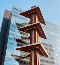 A modern architectural structure with three stacked platforms, each balanced on a central column. The platforms have a rust-colored finish, contrasting with the reflective glass facade of a high-rise building in the background. The image conveys an urban setting under a clear blue sky.