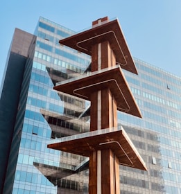 A modern architectural structure with three stacked platforms, each balanced on a central column. The platforms have a rust-colored finish, contrasting with the reflective glass facade of a high-rise building in the background. The image conveys an urban setting under a clear blue sky.