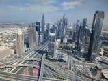 A bustling urban landscape featuring numerous modern skyscrapers and intricate highways, with clear skies overhead. The architecture is characterized by tall buildings with diverse designs and reflective glass windows. Roads and overpasses weave through the scene, highlighting an advanced infrastructure system.