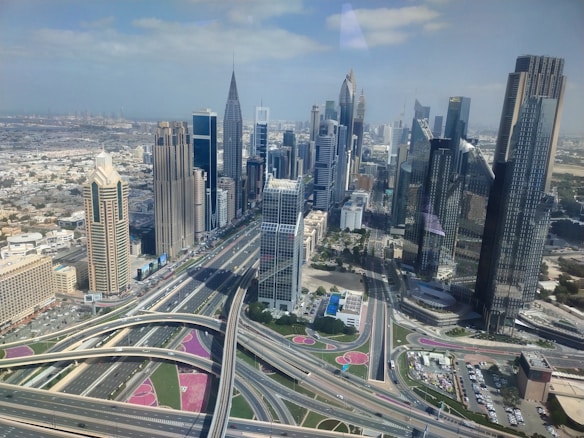 A bustling urban landscape featuring numerous modern skyscrapers and intricate highways, with clear skies overhead. The architecture is characterized by tall buildings with diverse designs and reflective glass windows. Roads and overpasses weave through the scene, highlighting an advanced infrastructure system.