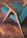 A modern wooden chair features teal fabric upholstery and angled wooden armrests, placed on a herringbone-patterned wooden floor. The wood has a natural finish, highlighting its grain. Metal rivets are visible on the chair's structure.