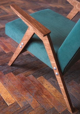 A modern wooden chair features teal fabric upholstery and angled wooden armrests, placed on a herringbone-patterned wooden floor. The wood has a natural finish, highlighting its grain. Metal rivets are visible on the chair's structure.