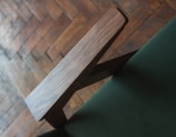 A close-up of a wooden chair armrest set against a green fabric, with a blurred wooden parquet floor in the background.
