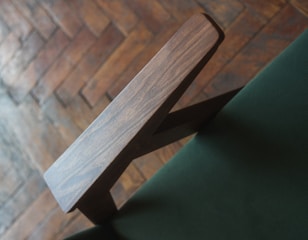 A close-up of a wooden chair armrest set against a green fabric, with a blurred wooden parquet floor in the background.