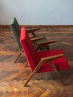 Two chairs with wooden frames and vibrant fabric upholstery, one in red and the other in green, are placed on a parquet wooden floor. The chairs have a minimalist, angular design and are positioned slightly at an angle from the side.
