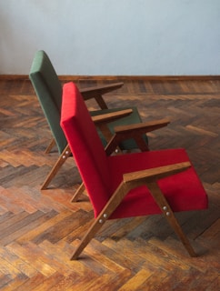 Two chairs with wooden frames and vibrant fabric upholstery, one in red and the other in green, are placed on a parquet wooden floor. The chairs have a minimalist, angular design and are positioned slightly at an angle from the side.