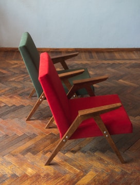 Two chairs with wooden frames and vibrant fabric upholstery, one in red and the other in green, are placed on a parquet wooden floor. The chairs have a minimalist, angular design and are positioned slightly at an angle from the side.