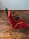 Two chairs with wooden frames and vibrant fabric upholstery, one in red and the other in green, are placed on a parquet wooden floor. The chairs have a minimalist, angular design and are positioned slightly at an angle from the side.